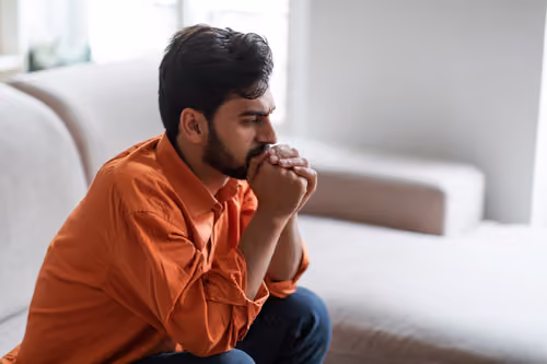 Older man sitting quietly with a thoughtful expression, hands folded, in a comfortable home setting.